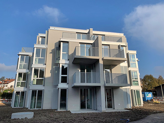 Ein modernes, mehrstöckiges Wohnhaus mit zahlreichen Balkonen und großen Fenstern. Die Fassade des Gebäudes besteht aus hellen Ziegeln, die Umgebung ist bebaut, im Vordergrund ist nackter Boden zu sehen. Der Himmel ist klar mit ein paar Wolken.