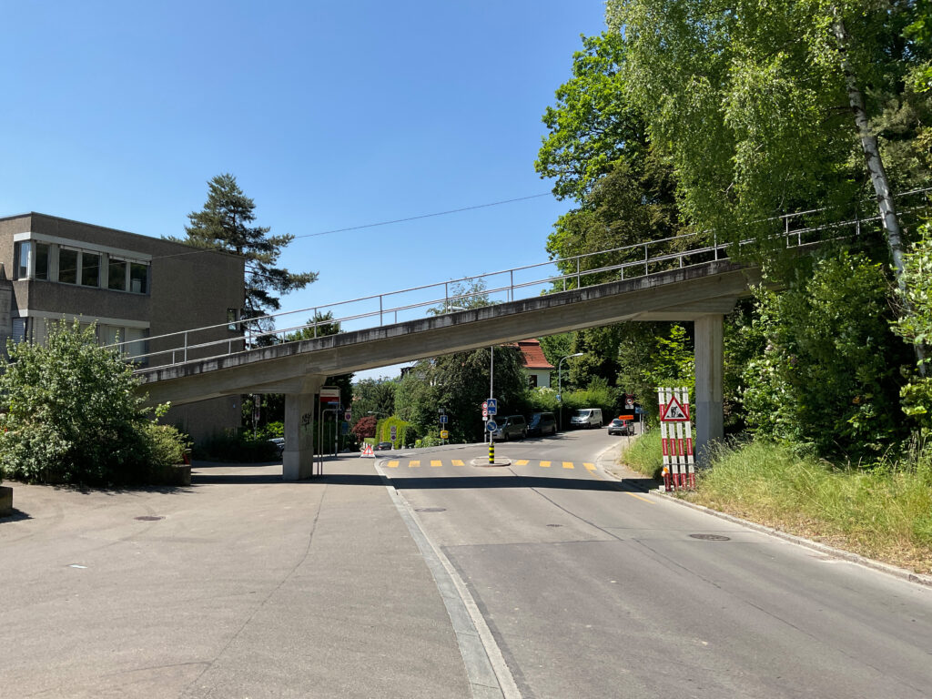 Eine ruhige Stadtstraße, die von einer Fußgängerbrücke durchquert wird, umgeben von Grünflächen und Gebäuden. Die Brücke hat Metallgeländer und darunter befinden sich Verkehrsschilder und ein kleiner Kreisverkehr. Auf beiden Seiten der Straße stehen Bäume unter einem klaren blauen Himmel.