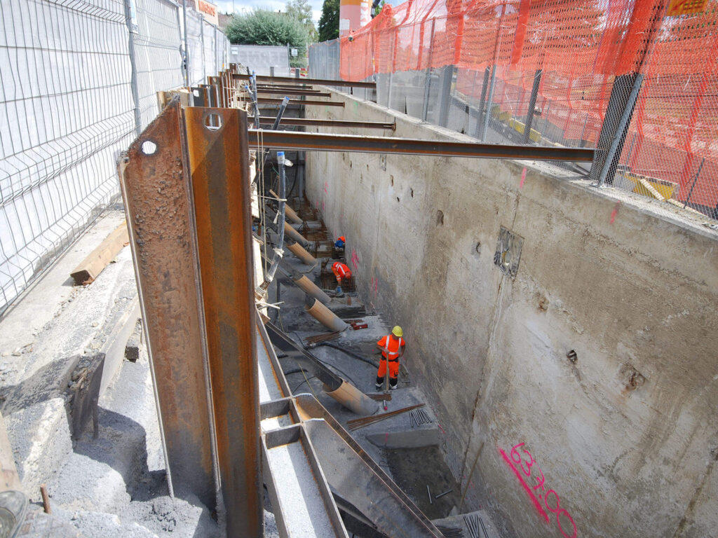 Ein Bauarbeiter in orangefarbener Warnkleidung und einem gelben Helm steht in einem großen, tiefen Betongraben. Der Graben wird von Metallträgern gestützt und oben ist ein orangefarbener Maschendrahtzaun angebracht. Überall auf der Baustelle sind verschiedene Baugeräte zu sehen.