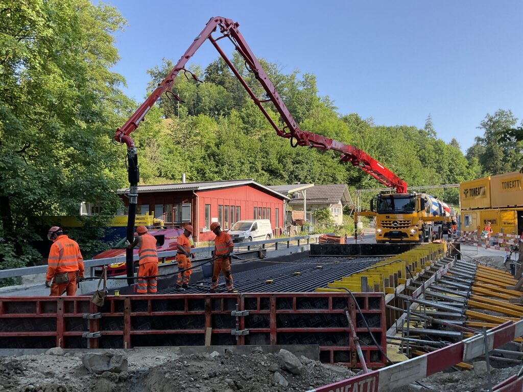 Baustelle mit mehreren Arbeitern in orangefarbener Schutzausrüstung, die einen Betonpumpenwagen bedienen und Beton in eine große Brückenfundamentschalung gießen. Die Baustelle ist von üppigen grünen Bäumen umgeben und umfasst ein kleines rotes Gebäude und Baugeräte.